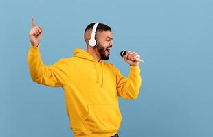 Joyful arab guy in headphones listening to music and singing song into microphone over blue studio background. Middle eastern man in headset enjoying karaoke photo