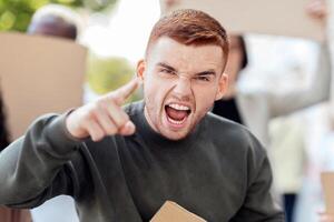 Charismatic young man leading multiracial group of protestors, pointing at camera and yelling something, standing over big international group of millennial people with empty boards, closeup photo
