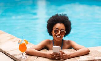 Joyful black girl with refreshing summer cocktail and smartphone relaxing in pool on sunny day, copy space photo