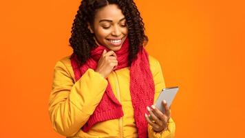 Black Woman With Mobile Phone Texting Reading Online Messages, Standing Holding Cellphone Over Yellow Studio Background, Wearing Winter Jacket. Smartphone Application Concept. Cropped, Copy Space photo