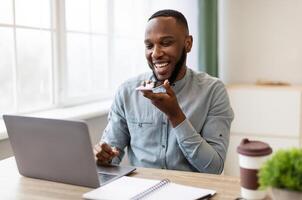 African Businessman With Smartphone Using Voice Assistant App Or Loudspeaker Talking Holding Phone Near Mouth Sitting At Workplace In Modern Office. IOT, Business Applications And Voice Search photo