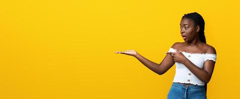 Surprized black woman pointing finger at invisible object at her open palm, standing over yellow background in studio, amazed african lady demonstrating free place for your mockup design, copy space photo