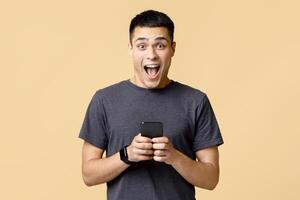 Great news about winning online or huge cashback. Happy excited shocking young man in casual with smart watch with open mouth hold smartphone, isolated on light sand background, empty space photo