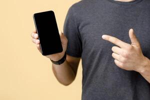 Best offer, ad and great review from blogger. Man in brown t-shirt recommends website and points finger at phone with blank screen, isolated on beige background, studio shot, close up, cropped photo