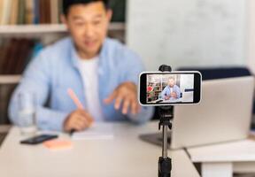 Educational Blogging. Asian Tutor Man Filming Online Lecture Via Smartphone Sitting At Workplace Indoor. E-Teaching Concept. Selective Focus On Mobile Phone photo