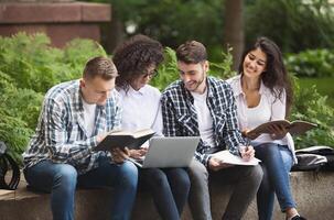 Multiethnic group of students sitting near college building, looking at laptop, reading books. Students before exam concept photo