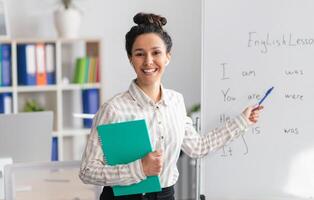 Training concept. Happy female tutor teaching English language, pointing at grammar rules on board and smiling at camera, holding clipboard in office interior photo