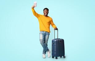 Cheerful african american millennial guy in casual outfit posing with suitcase, showing flight tickets and passport, having vacation, blue studio background, full length shot, copy space photo