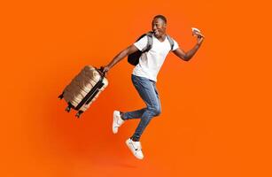 Bye-bye, Im going vacation. Young happy black man with suitcase, backpack, passport and flight tickets jumping up orange studio background. Excited african american guy running and looking back photo