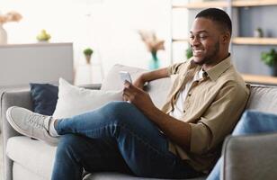 Great Mobile Application. Happy African American Man Using Phone Texting And Browsing Internet Sitting On Sofa At Home, Side View. Black Male Networking In Social Media Using App On Smartphone photo