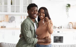 Excited african american young family showing keys to own home, happy black couple hugging, buying first house together, smiling husband and wife purchase new property. Ownership concept photo