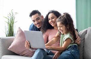 Cheerful arabic family having fun with laptop at home, watching photos together. Middle Eastern parents and their little daughter using computer while sitting on couch in living room, copy space