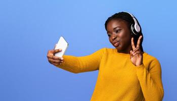 Cheerful Black Woman In Headphones Taking Selfie On Smartphone And Showing Peace Gesture While Posing At Camera, Joyful African Lady Having Fun, Posing Over Purple Background In Studio, Copy Space photo