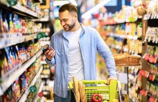 Consumption Concept. Portrait Of Smiling Bearded Guy With Shopping Cart In Market Buying Groceries Food Walking Along The Aisle And Shelves In Store, Holding Glass Jar Of Sauce, Checking Label photo