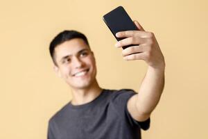 Happy blogger makes streaming or photo for social media. Smiling generation z man in brown t-shirt makes selfie, blurry and focus on smartphone, isolated on beige background, free space