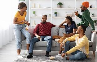 Group Of Black Friends Enjoying Drinks Party At Home On Weekend. Cheerful african american young men and women sitting in living room, drinking beer and having conversation, home party concept photo