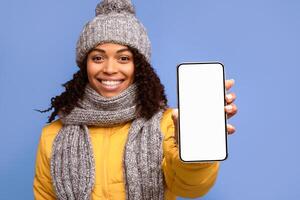 African Woman Showing Smartphone Empty Screen Recommending Mobile Phone Application Posing Wearing Winter Jacket Standing Over Purple Studio Background. Cellphone Display Mockup Advertisement photo