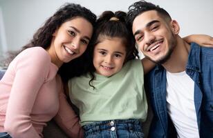 Closeup Portraif Of Happy Arabic Family Of Three With Little Daughter, Cheerful Middle Eastern Parents Posing At Camera While Having Fun With Their Little Child At Home, Relaxing On Couch Together photo