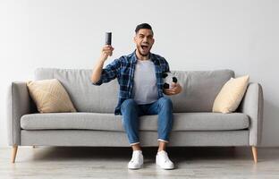 Portrait of excited young guy watching football match, raising clenched fist. Emotional man sitting on couch cheering favorite team enjoying game goal on TV at home holding remote controller and ball photo