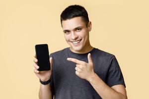 Excited winner man demonstrating mobile phone after received good news. Smiling young guy, points finger at smartphone with blank screen, advising good device, isolated on beige background photo