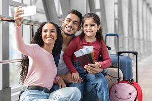 Airport Selfie. Happy Middle Eastern Family Of Three Taking Photo With Smartphone While Waiting For Flight At Terminal, Cheerful Arabic Parents And Daughter Smiling And Posing At Camera, Free Space