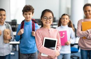 Pupils, Technology And Learning Concept. Smiling asian girl in eyeglasses holding digital tablet and standing with group of multicultural classmates in the background, studying at elementary school photo