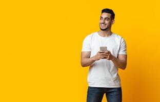 Cheerful arab guy with smartphone looking at copy space and smiling, yellow studio background, panorama. Positive middle-eastern young man chatting on mobile phone, using new application photo