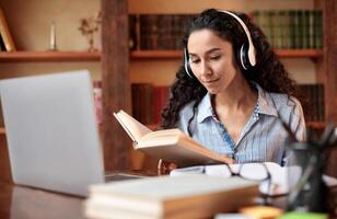 Portrait of calm young woman wearing wireless headset, reading paper book, sitting at table with laptop computer. Lady studying online at home, doing homework, enjoying story, preparing for exam photo