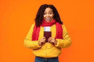 Joyful African Tourist Lady Holding Tickets And Passport Going On Winter Vacation Posing Over Yellow Studio Background, Smiling To Camera. Traveling On Christmas Holidays, Tourism Concept. photo
