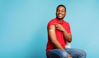 Vaccinated African Guy Showing Arm After Coronavirus Vaccination And Getting Covid-19 Vaccine Injection Sitting On Blue Background. Flu And Covid Virus Prevention. Panorama, Empty Space photo