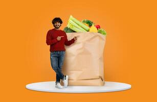 A smiling shopper points at a large paper bag overflowing with fresh vegetables and fruits. The vibrant orange background highlights the new normal of grocery shopping at home. photo