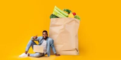 A cheerful customer sits next to a large paper bag filled with fresh groceries while using a laptop to order food. This reflects the new normal of online shopping. photo