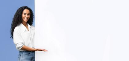 Place For Your Ad. Smiling brunette woman pointing at white advertisement board with open palm, demonstrating copy space for your text or design, standing over yellow studio background, panorama photo