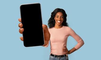 App Ad. Portrait of smiling African American woman holding smartphone with black blank device screen in hand close up to camera, blue studio. Gadget with empty free space for mock up, banner isolated photo