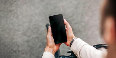 Social networks, work online, good app and modern lifestyle. Young man freelancer or blogger sits on bicycle and looks in smartphone with blank screen in hands, outdoor, cropped, free space photo