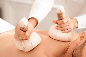 Closeup of compress balls in unrecognizable masseuse hands making herbal massage for handsome bearded man. Middle-aged man spending day at male spa, getting healing herbal massage photo