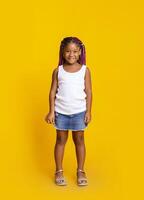 Portrait of cute little black girl with afro braids smiling and looking at camera while standing in studio over yellow background, copy space photo