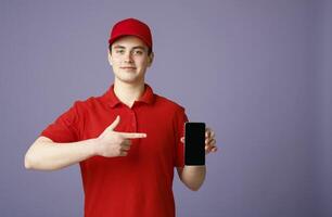 Timely delivery. Cute courier in red uniform points finger at smartphone with blank screen, isolated gray background photo