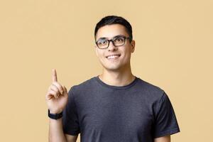Smart student has idea or sees cool advertisement. Cute smiling young man blogger in glasses, points with finger and looks up at free space, isolated on beige background, studio shot, free space photo