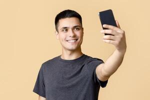 Modern photo on gadget for blog. Cheerful young man student makes selfie on phone for social networks and streaming with advises, isolated on beige background, studio shot, free space