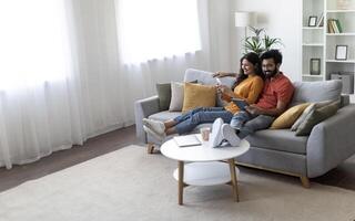 Happy Young Indian Spouses Relaxing With Modern Gadgets On Couch At Home, Married Eastern Couple Resting In Living Room Interior, Man Using Digital Tablet While Woman Browsing App On Smartphone photo