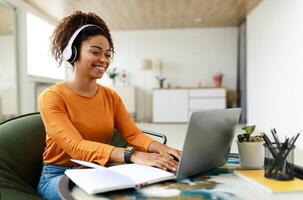 People And Technology. Portrait of young smiling black woman in wireless headset using pc sitting at table on chair in living room, typing on keyboard. Cheerful lady browsing internet, surfing web photo