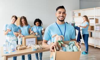 Arab male volunteer holding box with clothes and toys for donation, smiling to camera while working at charity center with group of colleagues, packing products in boxes on background photo
