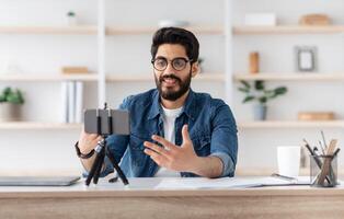 Arab male blogger recording at home with smartphone on tripod, middle-eastern man making content for his blog, sitting at home office and gesturing at phone camera, copy space photo