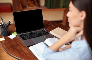 E-learning Concept. Over the shoulder view of woman sitting at desk with open notebook, using laptop with black empty screen for mockup template, watching webinar, studying online at home photo