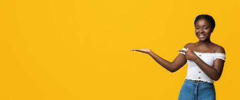 Happy Young African American Woman Pointing Finger At Open Palm, Posing Over Yellow Background In Studio, Demonstrating And Advertising Something, Holding Invisible Object, Panorama With Copy Space photo