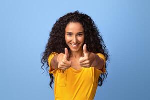Joyful brunette woman gesturing thumbs up at camera, demonstrating sign of approval, recommending something, positive millennial lady standing over purple background in studio, copy space photo