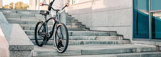 A guy navigates down a set of concrete stairs with his bike, showcasing an active lifestyle in a modern urban setting during the daytime. The scene emphasizes movement and energy, cropped photo