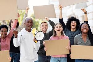 Furious multiracial students with empty placards having strike or demonstration on the street. Motivated african american guy with loudspeaker chanting slogans over demonstrating crowd outdoors photo