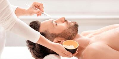 Side view of handsome caucasian middle-aged man getting face treatment at spa, closeup. Unrecognizable beautician applying detox white mask on relaxed man face, using brush and clay photo